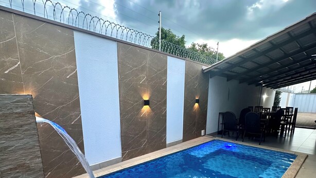 A swimming pool with a water feature, surrounded by a wall with light fixtures and a covered area with chairs. - Davisis Luxo Piscina Prox Graciosa (Palmas)