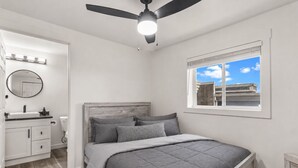 A bedroom with a bed, a ceiling fan, a window with a view, and a bathroom with a sink and mirror.