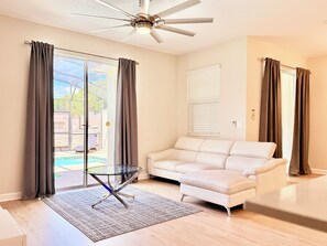 Ein modernes Wohnzimmer mit Glastisch, weißem Sofa, Deckenventilator und Blick auf einen Swimmingpool.