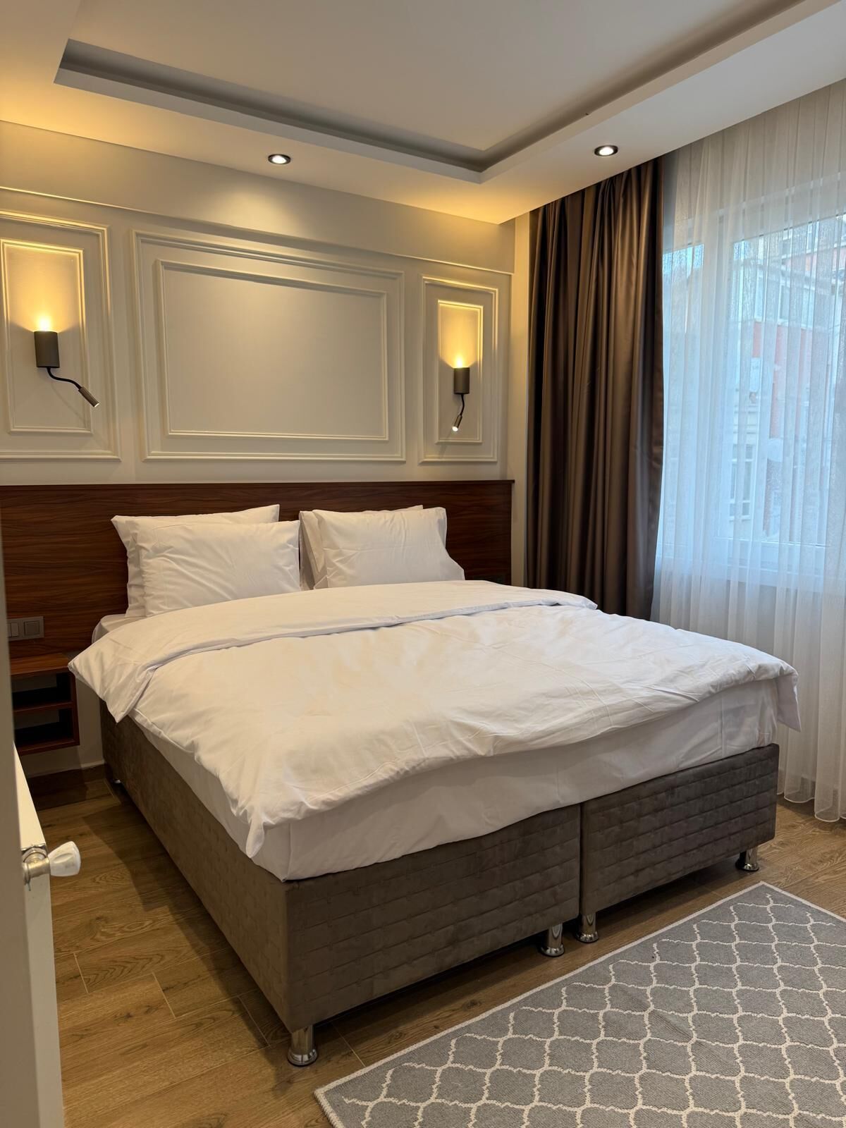 A hotel room with a large bed, two wall-mounted lights, a wooden headboard, and a window with curtains.