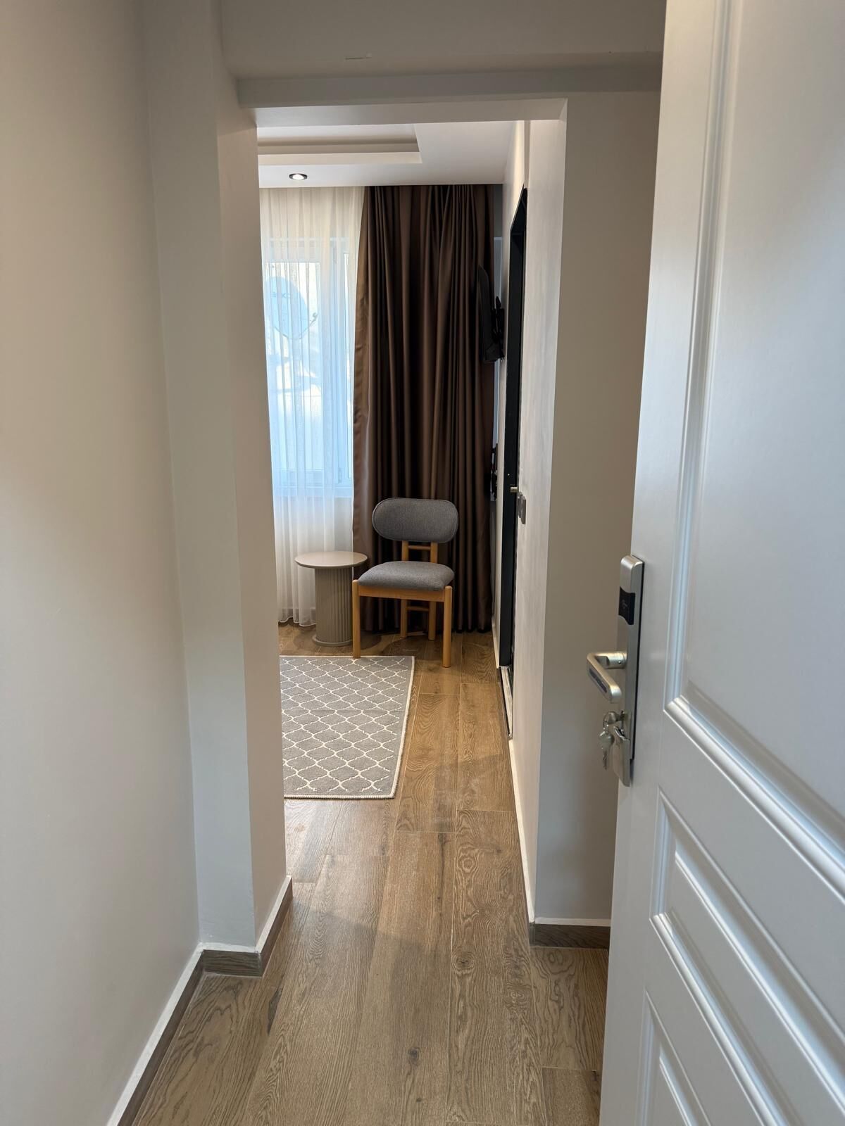 A hallway leading to a room with a wooden floor, a grey chair, a small table, and a window with curtains.