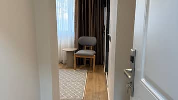 A hallway leading to a room with a wooden floor, a grey chair, a small table, and a window with curtains.