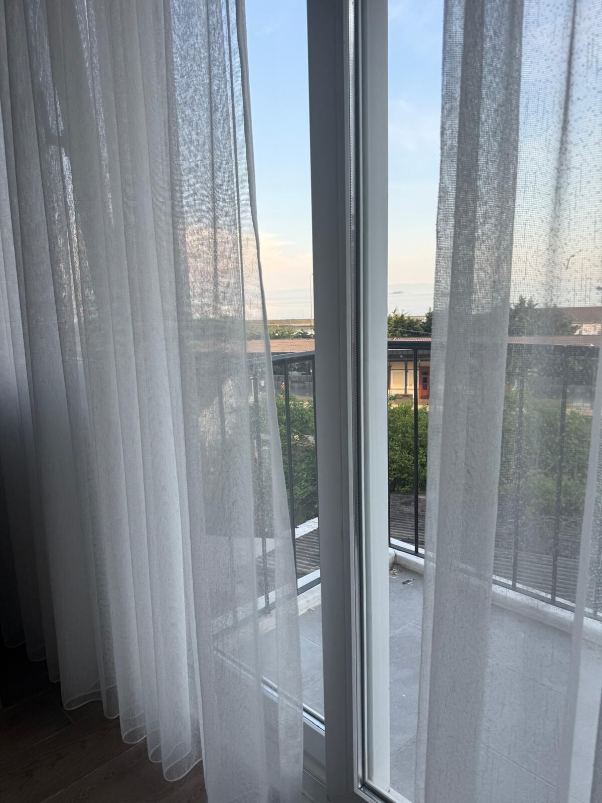 A room with sheer curtains, a large window, and a view of a building and greenery.