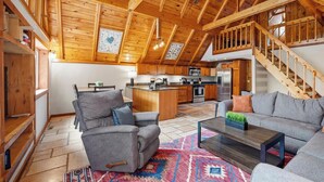 Ein geräumiges Wohnzimmer mit hoher Decke, Holzbalken, einem grauen Sofa, einem Couchtisch und einer Küche im Hintergrund.