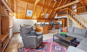 A spacious living room with a high ceiling, wooden beams, a gray sofa, a coffee table, and a kitchen in the background. - At Pond View, you can fish from the pond, soak in the hot tub, gather around the fire. (Twin Lakes)