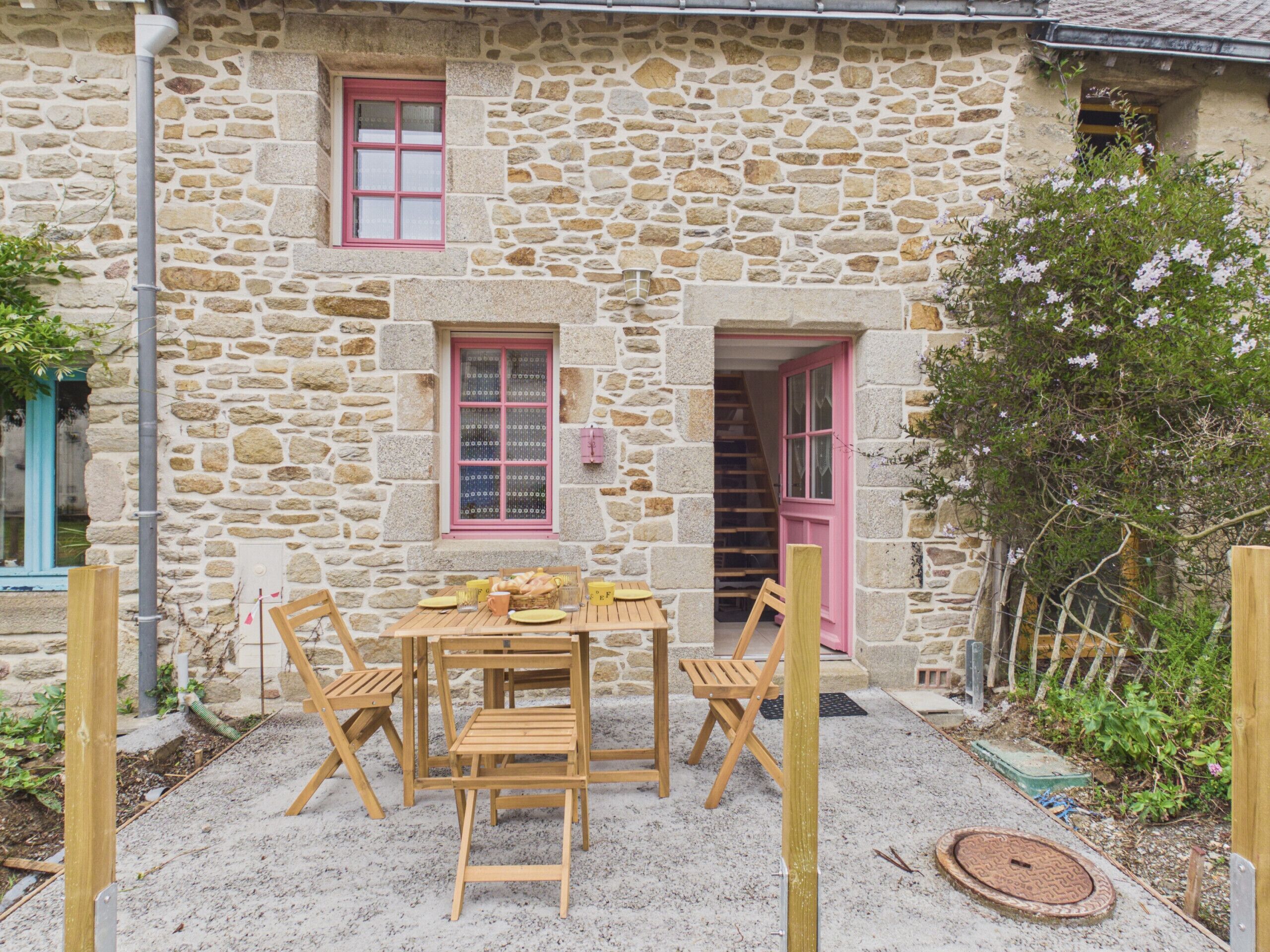 A stone building with a wooden table and chairs, a pink door, and a window with pink shutters.