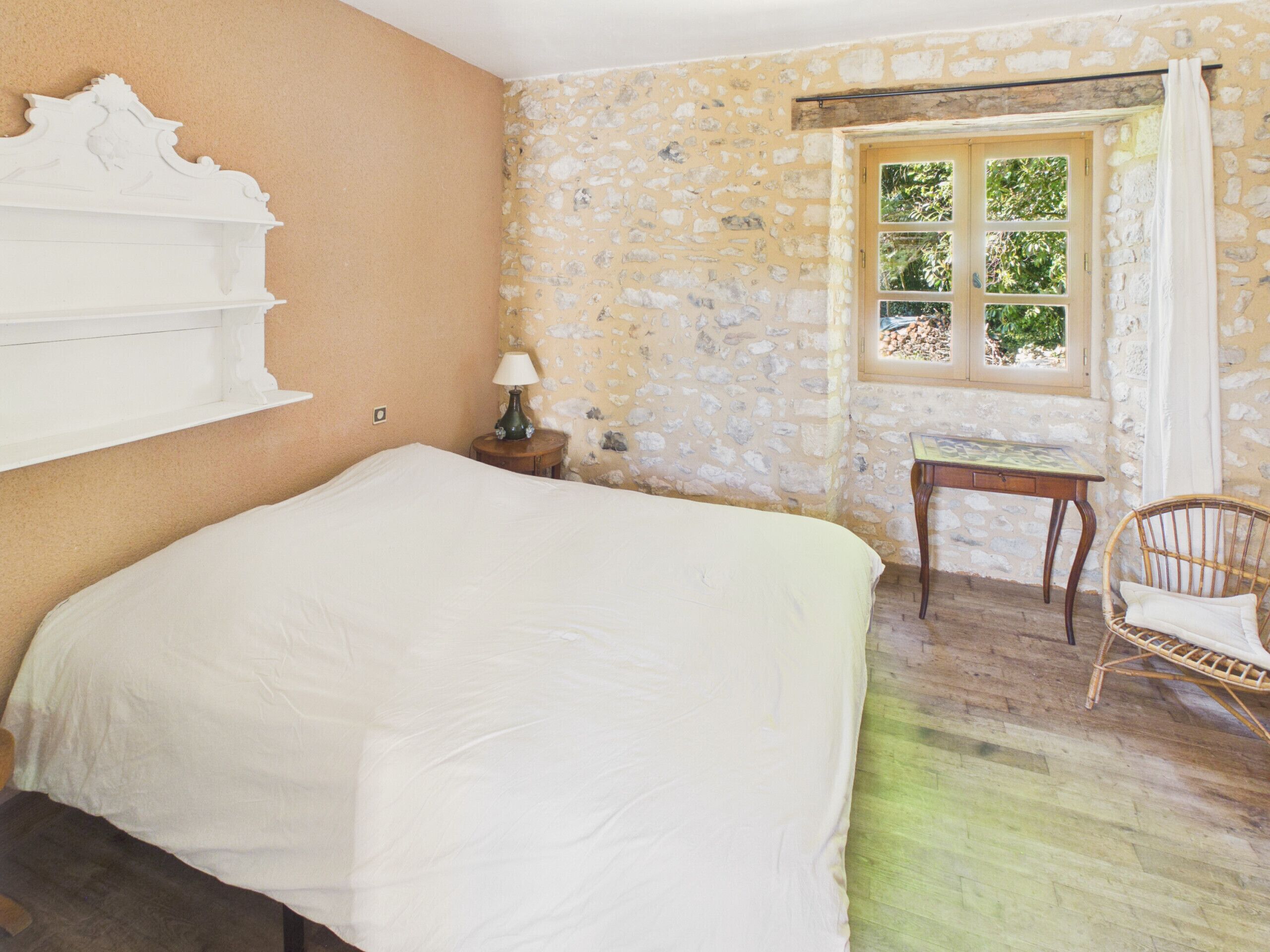 Une chambre à coucher avec un lit, une petite table, une chaise, une fenêtre avec des rideaux et un mur en pierre.