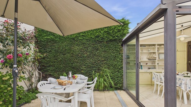 Een terras buiten met een eettafel, omringd door witte stoelen, een grote parasol en een glazen constructie met uitzicht op het interieur.