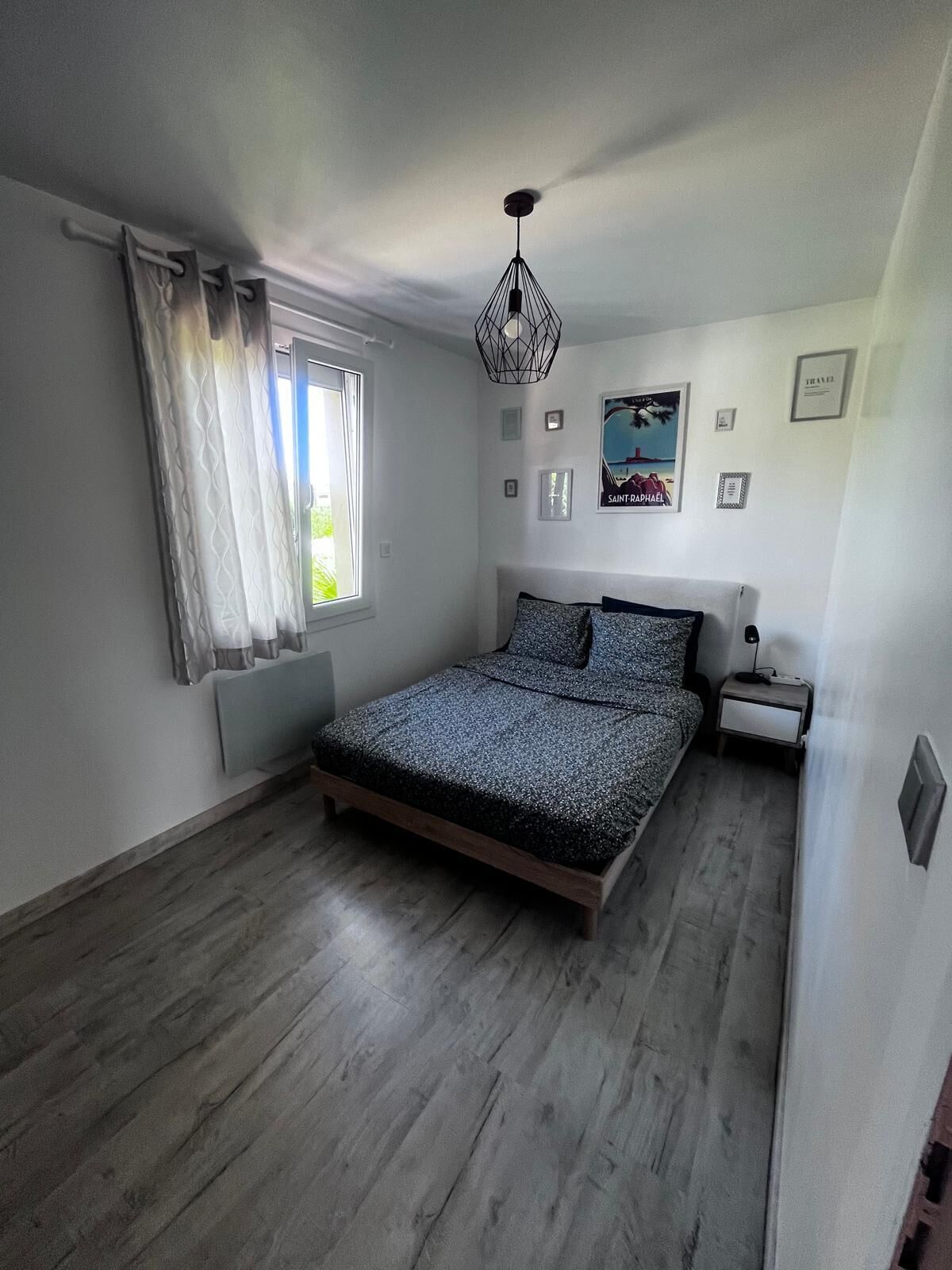 Une chambre à coucher comprenant un lit, une table de chevet, une fenêtre avec des rideaux, un luminaire fixé au mur et un tableau encadré.