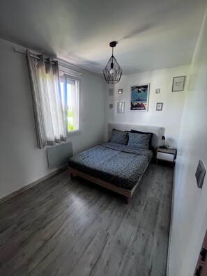 Een slaapkamer met een bed, nachtkastje, raam met gordijnen, een wandlamp en een lijst met een afbeelding.