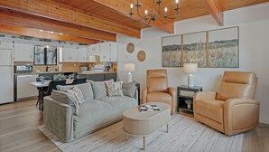 En moderne stue med sofa, sofabord og to læderstole, et køkken med hvide skabe, og et spiseområde.