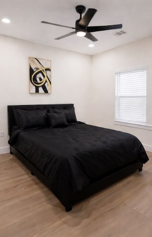 Quarto com cama preta, ventoinha no teto, janela com persianas e uma obra de arte abstrata na parede.