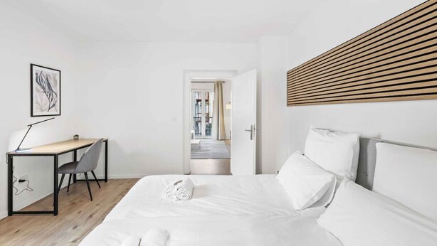 Et moderne hotelværelse med seng, skrivebord og stol, samt en dør der fører til en balkon og en sengegavl af trælameller.