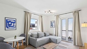 En moderne stue med en grå hjørnesofa, et rundt sofabord og et lille rundt bord med en stol. Der er to vinduer med gardiner, en loftlampe og kunst på væggene.