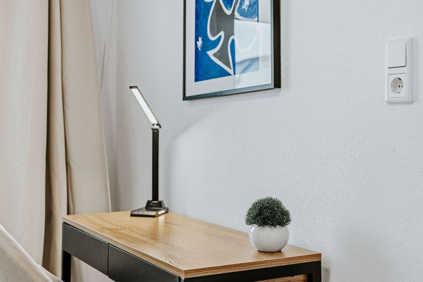 Un bureau en bois, une lampe de bureau noire, un vase blanc contenant une petite plante et, au mur, un tableau abstrait encadré.