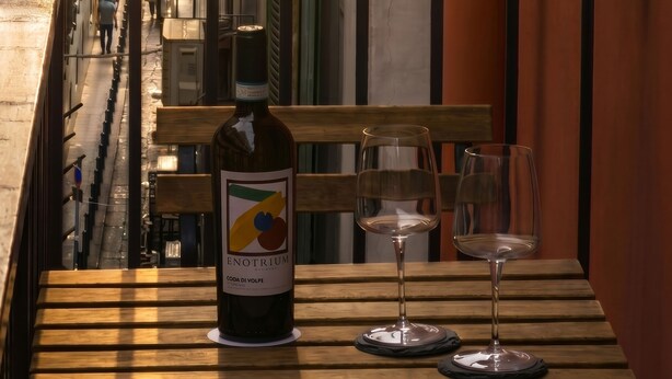 Een balkon met een fles wijn en twee glazen op een houten tafel, met uitzicht op een smalle straat met gebouwen.