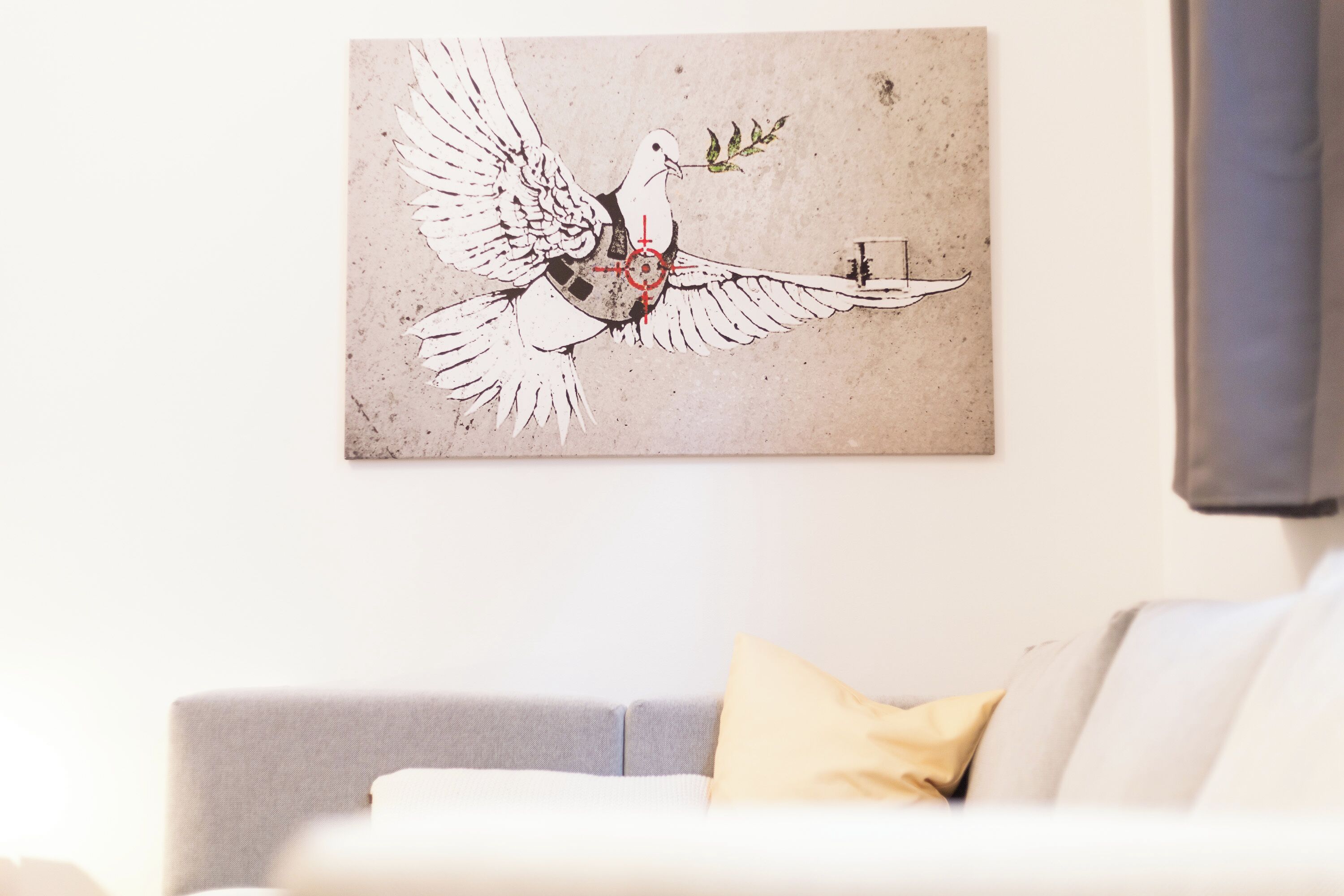 A wall-mounted artwork of a dove with a cross, a grey sofa, and a yellow pillow.