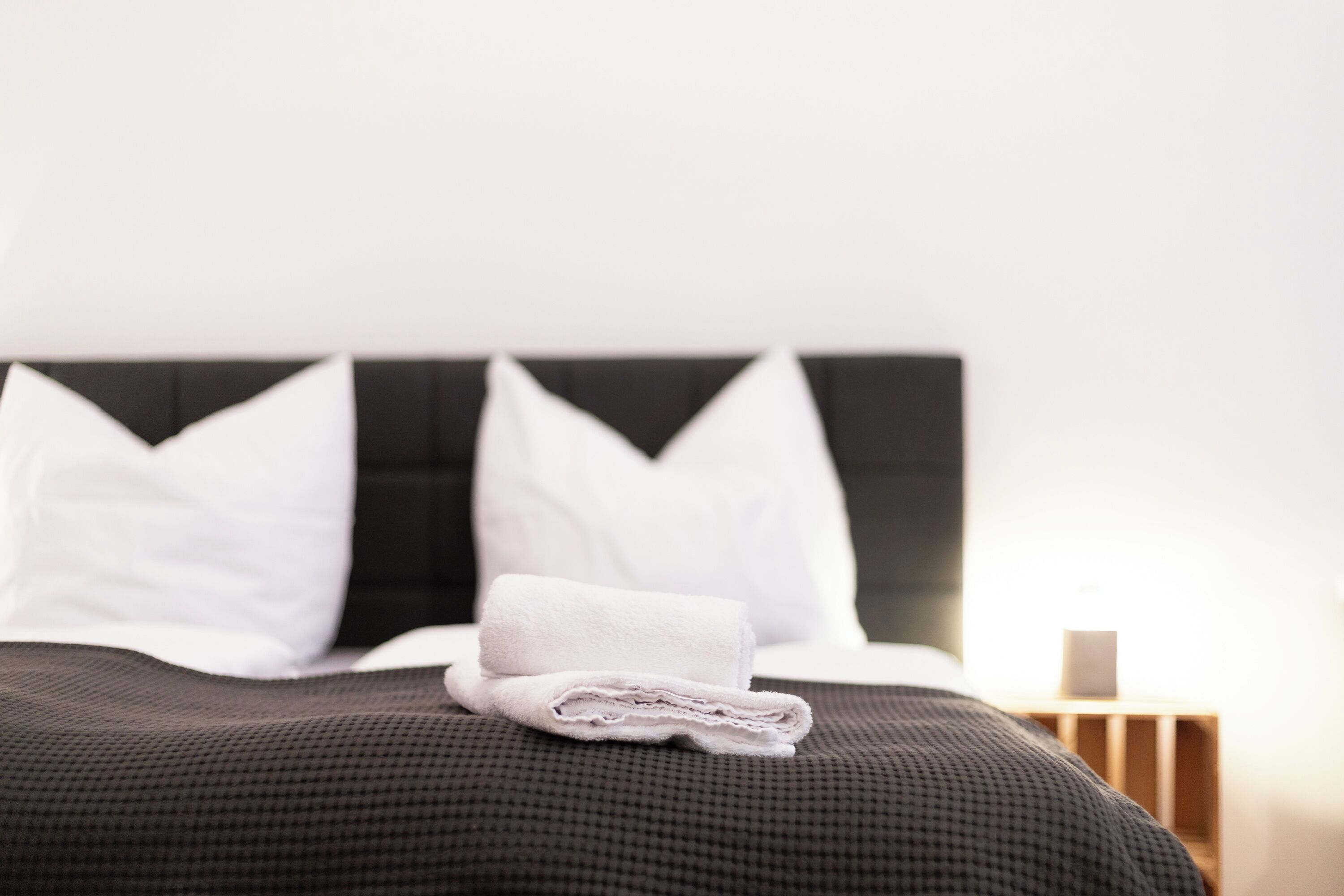 Ein ordentlich bezogenes Bett mit einem gefalteten weißen Handtuch, einem schwarzen Kopfteil und weißen Kissen.