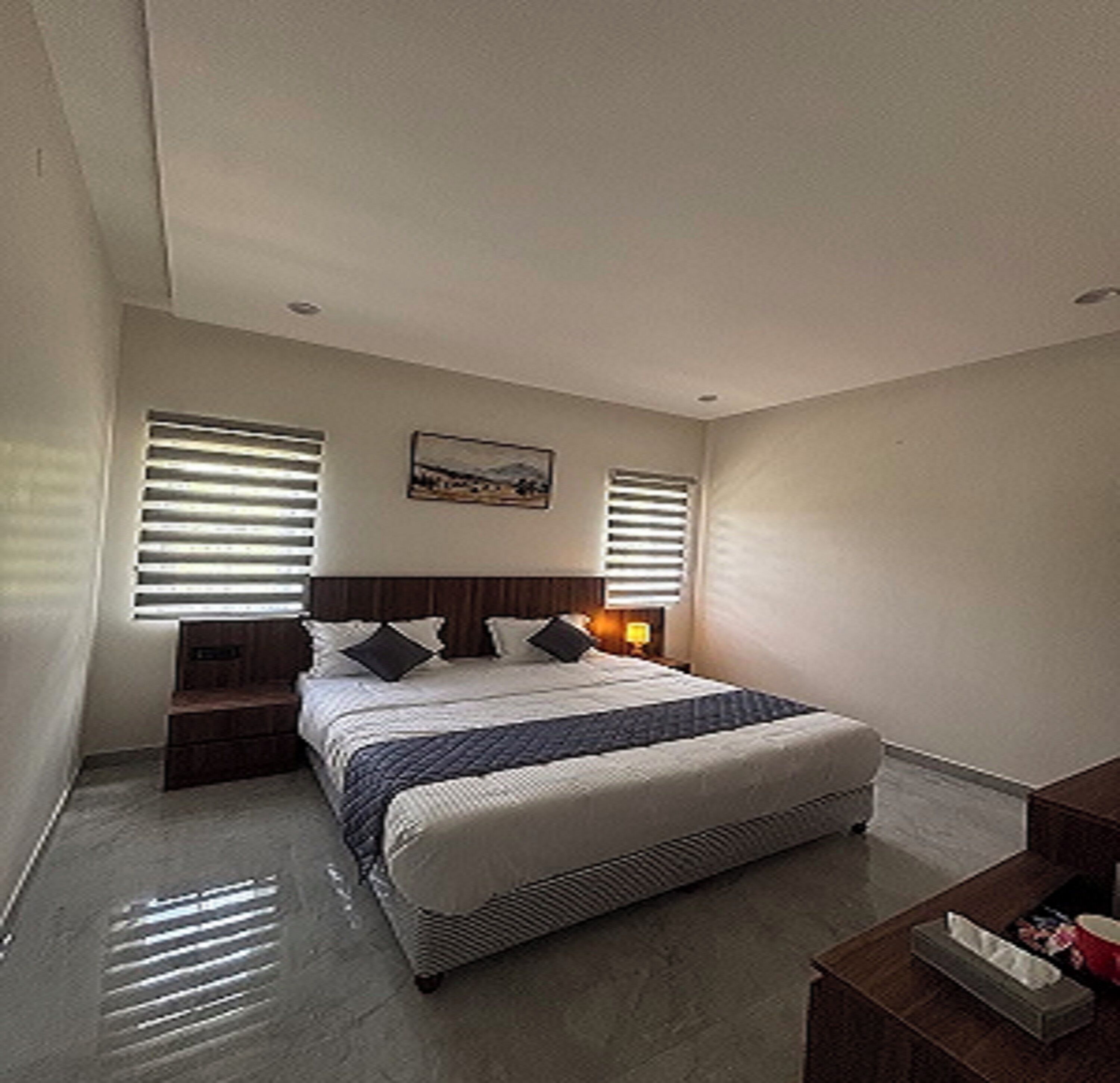 A bedroom with a bed, two nightstands, a bedside table, a window with blinds, and a wall-mounted picture.