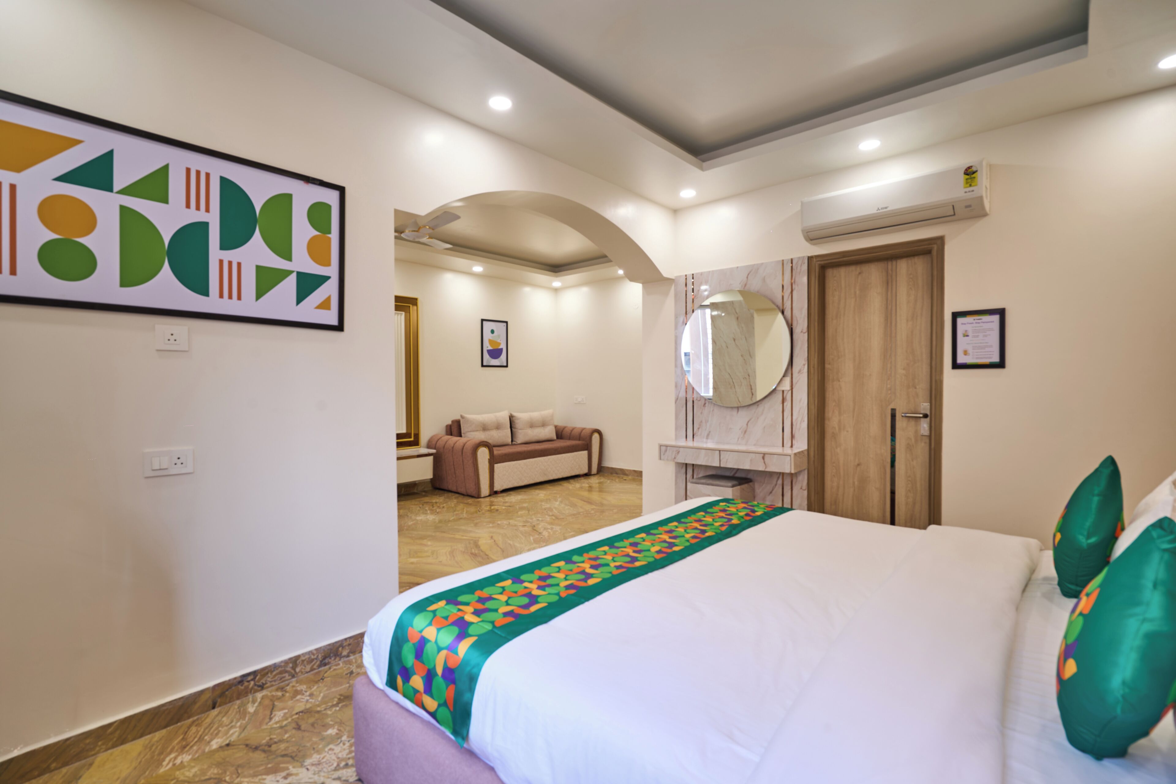 A hotel room with a large bed, a sofa, a mirror, and a wall art piece.