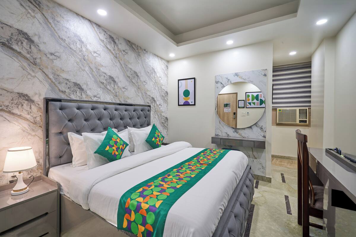 Un dormitorio con pared de mármol, cabecera capitonada, cama con ropa de cama blanca, manta estampada verde, mesita de noche con lámpara y escritorio con silla.