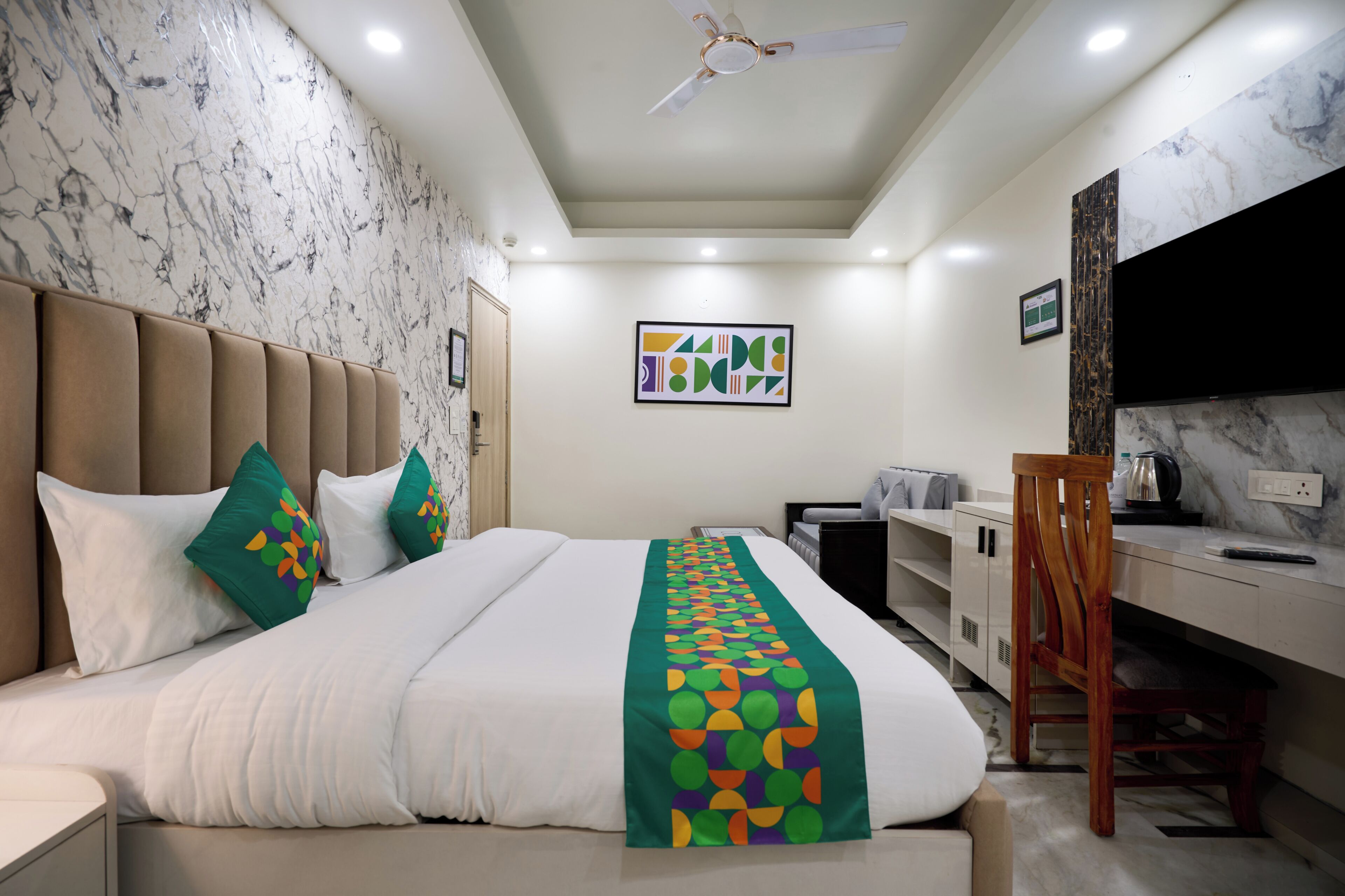 A hotel room with a bed, a desk, a chair, a ceiling fan, and a wall-mounted TV.