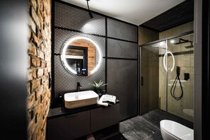 Une salle de bain moderne avec un mur en briques, un miroir rond, un lavabo blanc et une douche Ă parois de verre.