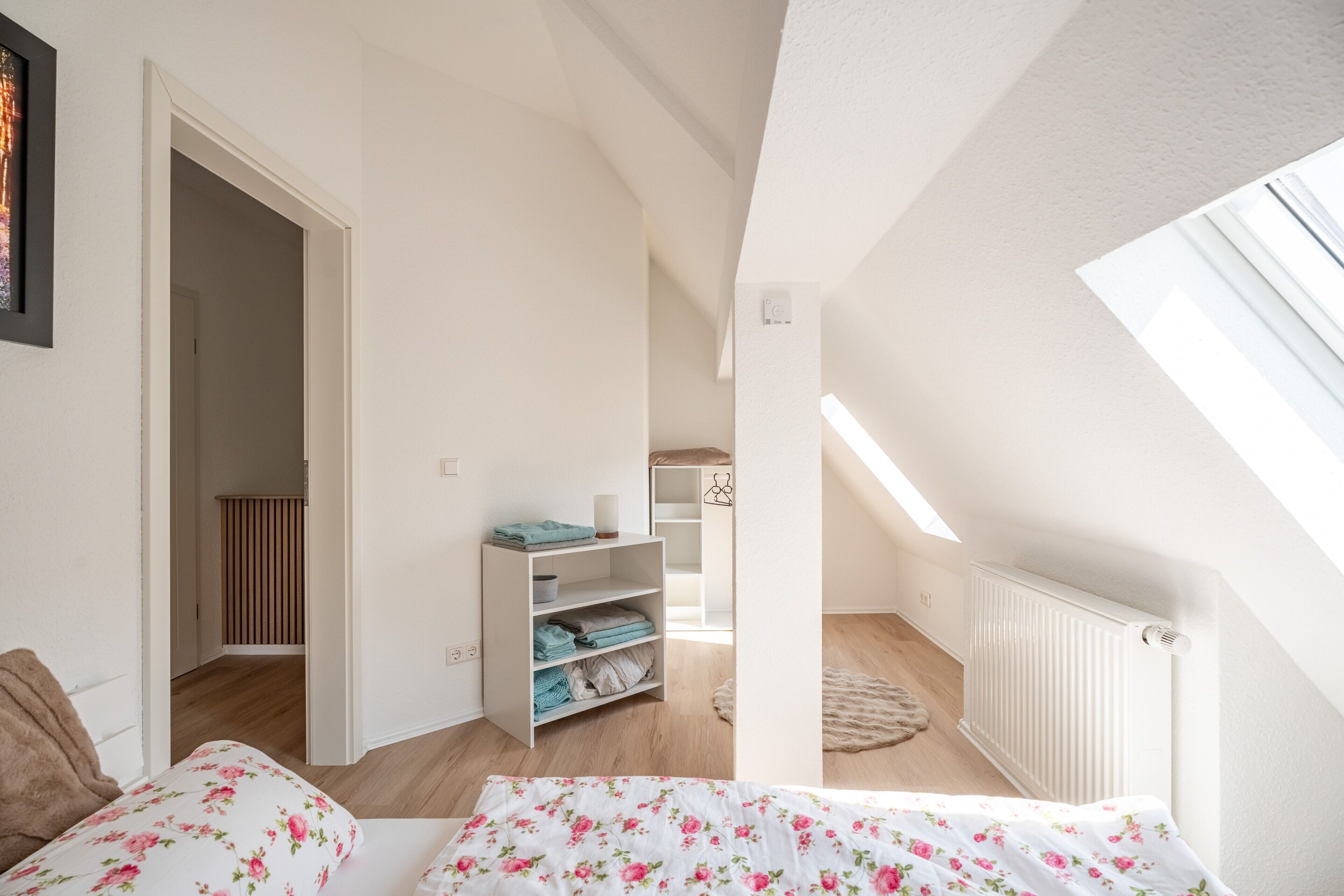A bedroom with a skylight, a bed with floral bedding, a white wardrobe, and a radiator.