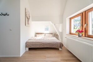 Une chambre avec un plafond en pente, un lit avec une literie à motifs floraux, une fenêtre donnant sur un beau paysage et un radiateur.