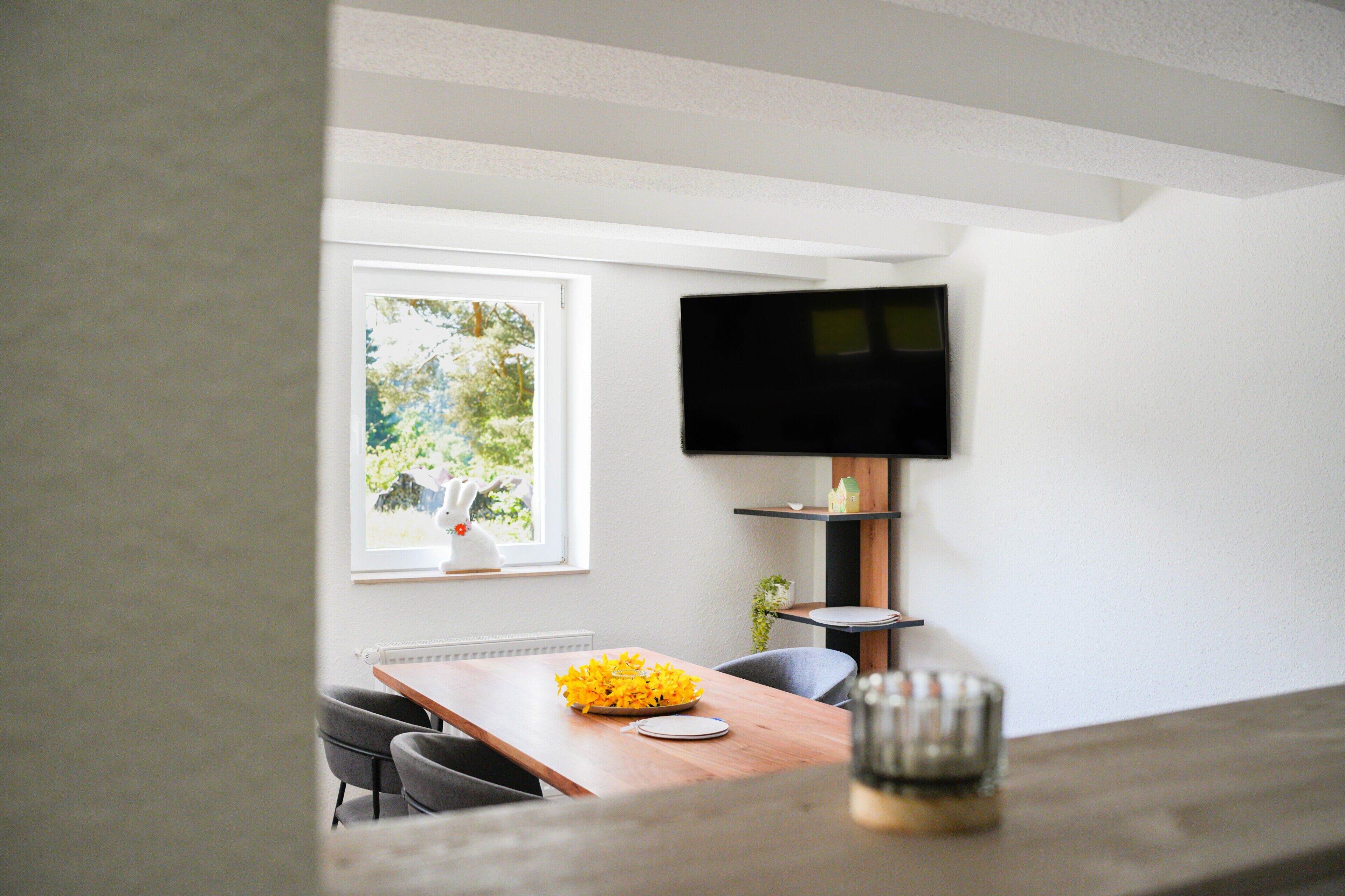 A dining area with a wooden table, chairs, a bowl of yellow flowers, a television mounted on the wall, and a window with a view of greenery.