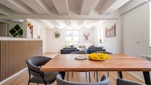 Un espace repas moderne avec une table en bois et des chaises grises, une cuisine en arrière-plan et une vue sur l’extérieur grâce à une grande fenêtre.