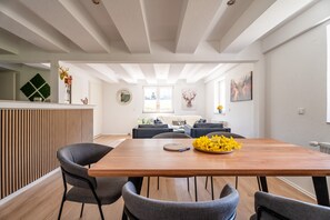 Ein moderner Essbereich mit einem Holztisch und grauen Stühlen, im Hintergrund eine Küche und durch ein großes Fenster Blick ins Freie.