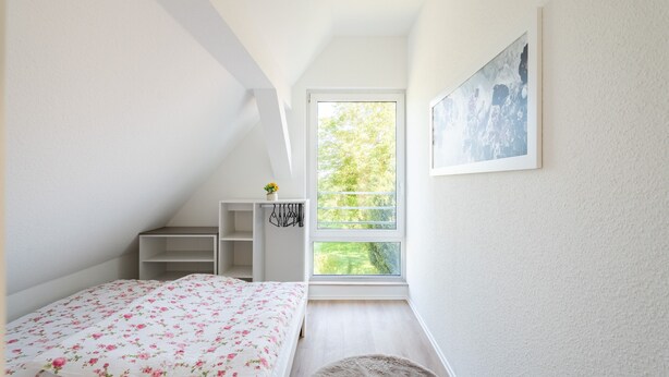 Ein Schlafzimmer mit Bett, Fenster, einem Gemälde und einer kleinen Ablage mit einer Pflanze.