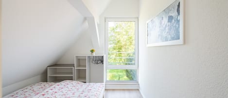 A bedroom with a bed, a window, a painting, and a small shelf with a plant.