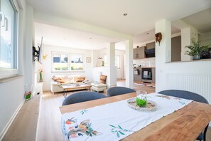 A modern living room with a dining table, sofas, and a kitchen in the background.