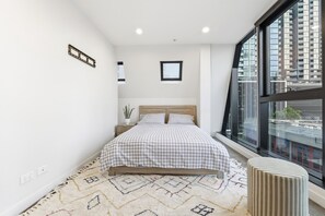 Et moderne soveværelse med en stor seng, et vindue med byudsigt, et natbord og en lille skammel.