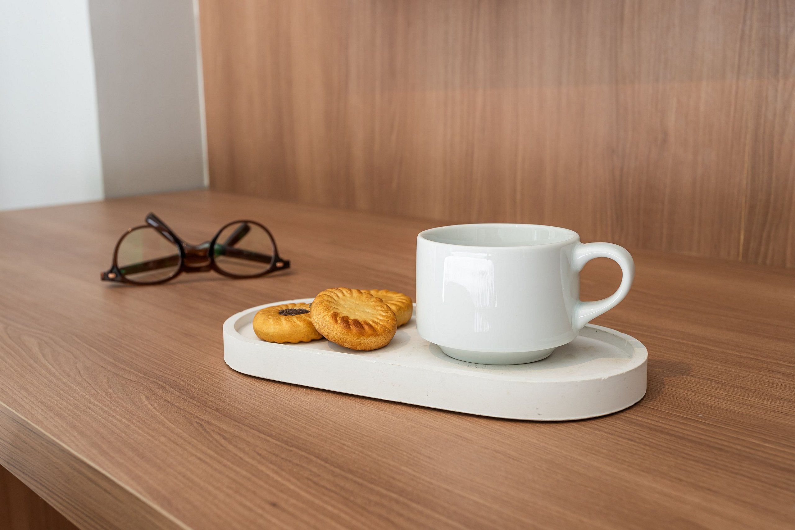 A white mug on a coaster, a pair of glasses, and two pastries on a wooden surface.