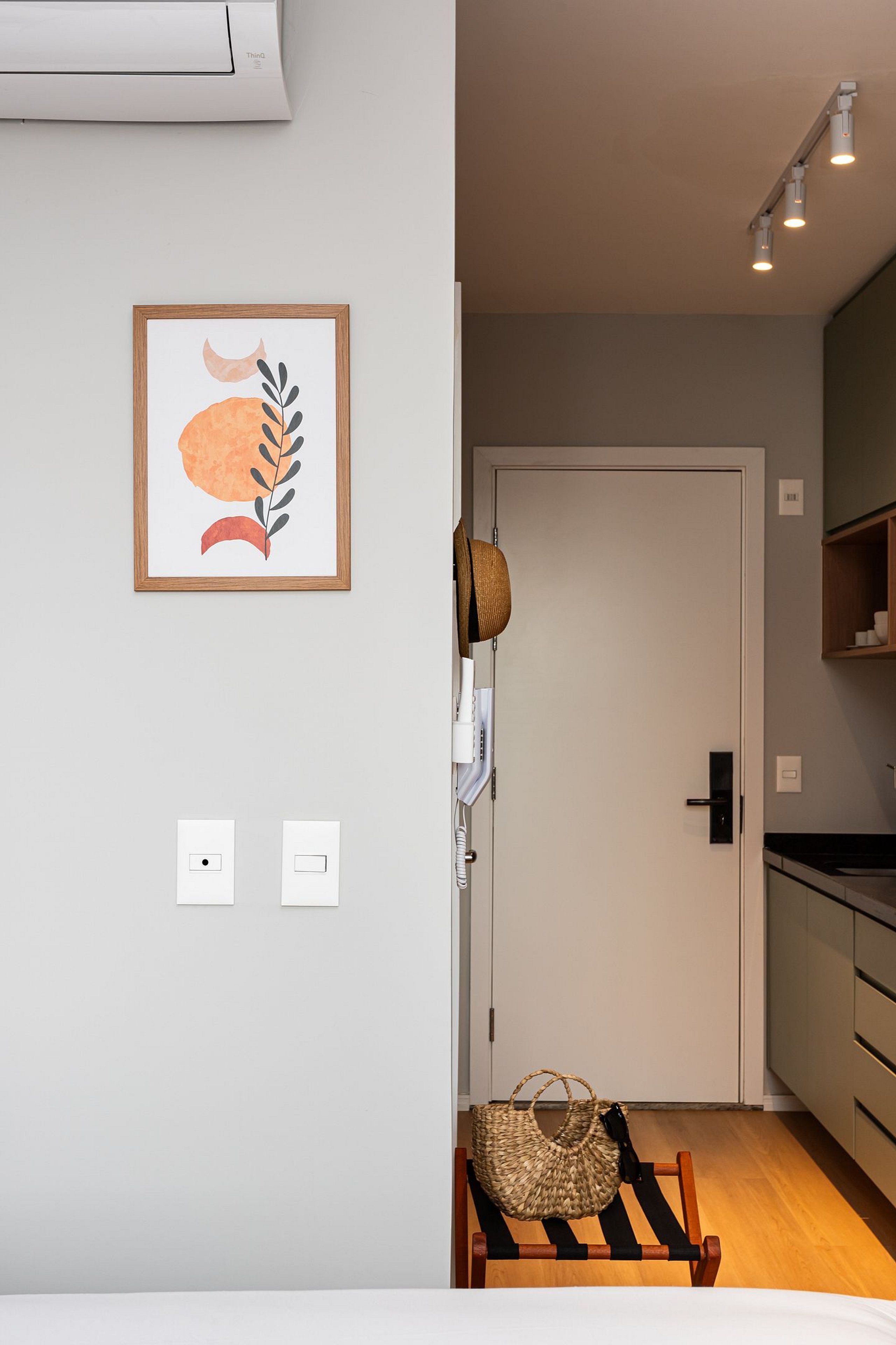 Une pièce comprenant un lit, une porte, un luminaire fixé au mur, un tableau encadré et un panier en osier.