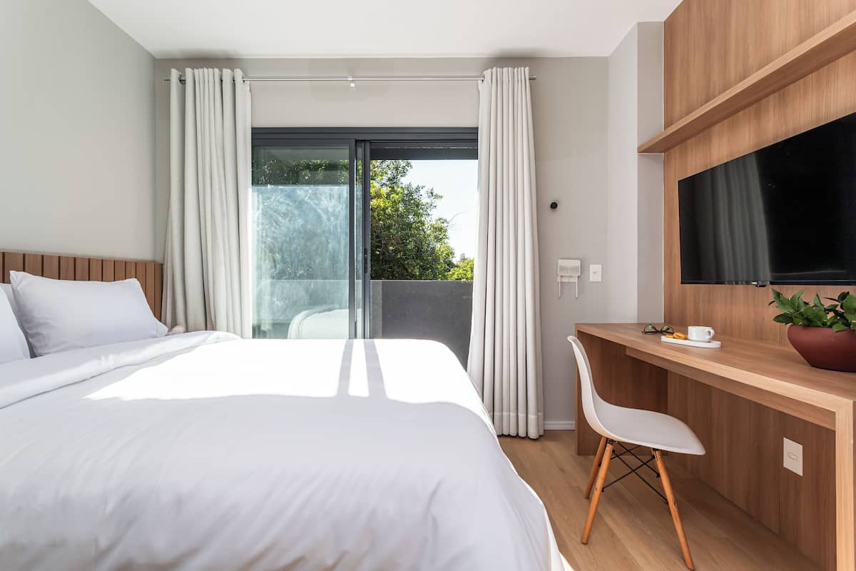Habitación de hotel moderna con una cama grande, un escritorio, una silla, un televisor y un balcón con cortinas.