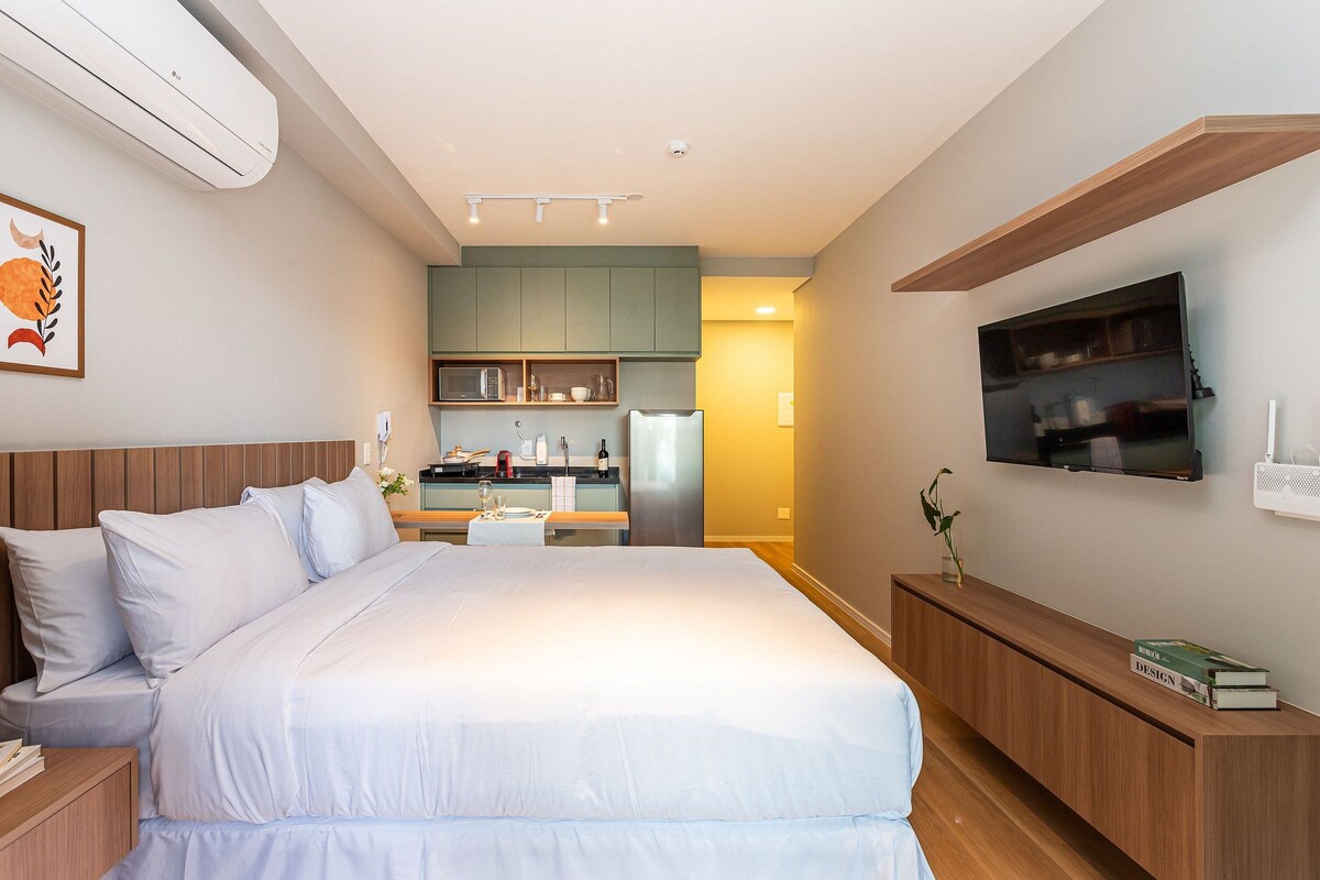 Habitación de hotel moderna con una cama grande, un televisor de pantalla plana, una mesita de noche de madera y una kitchenette al fondo.