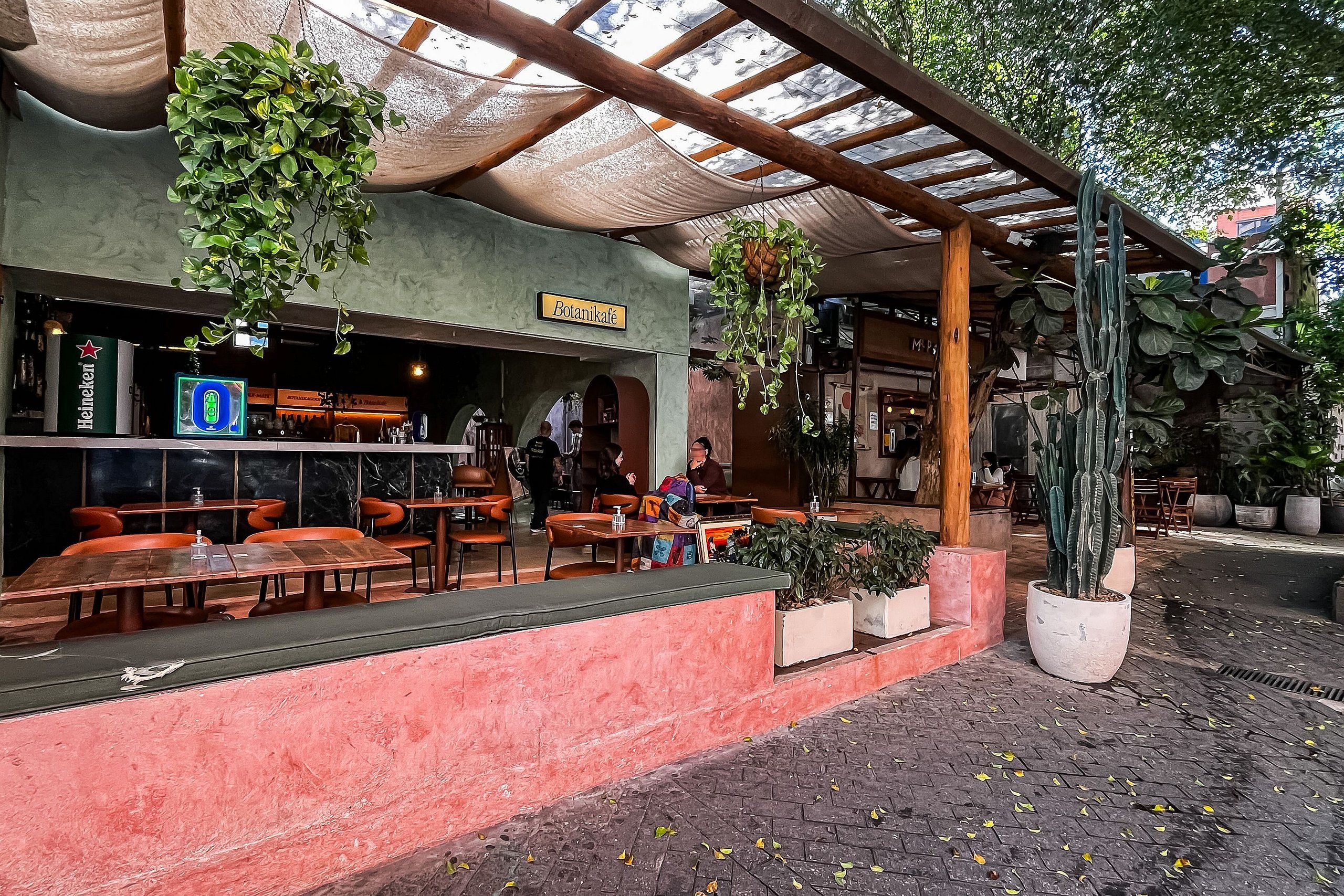 An outdoor café with tables and chairs, a sign reading 'Botanikafé', and hanging plants.