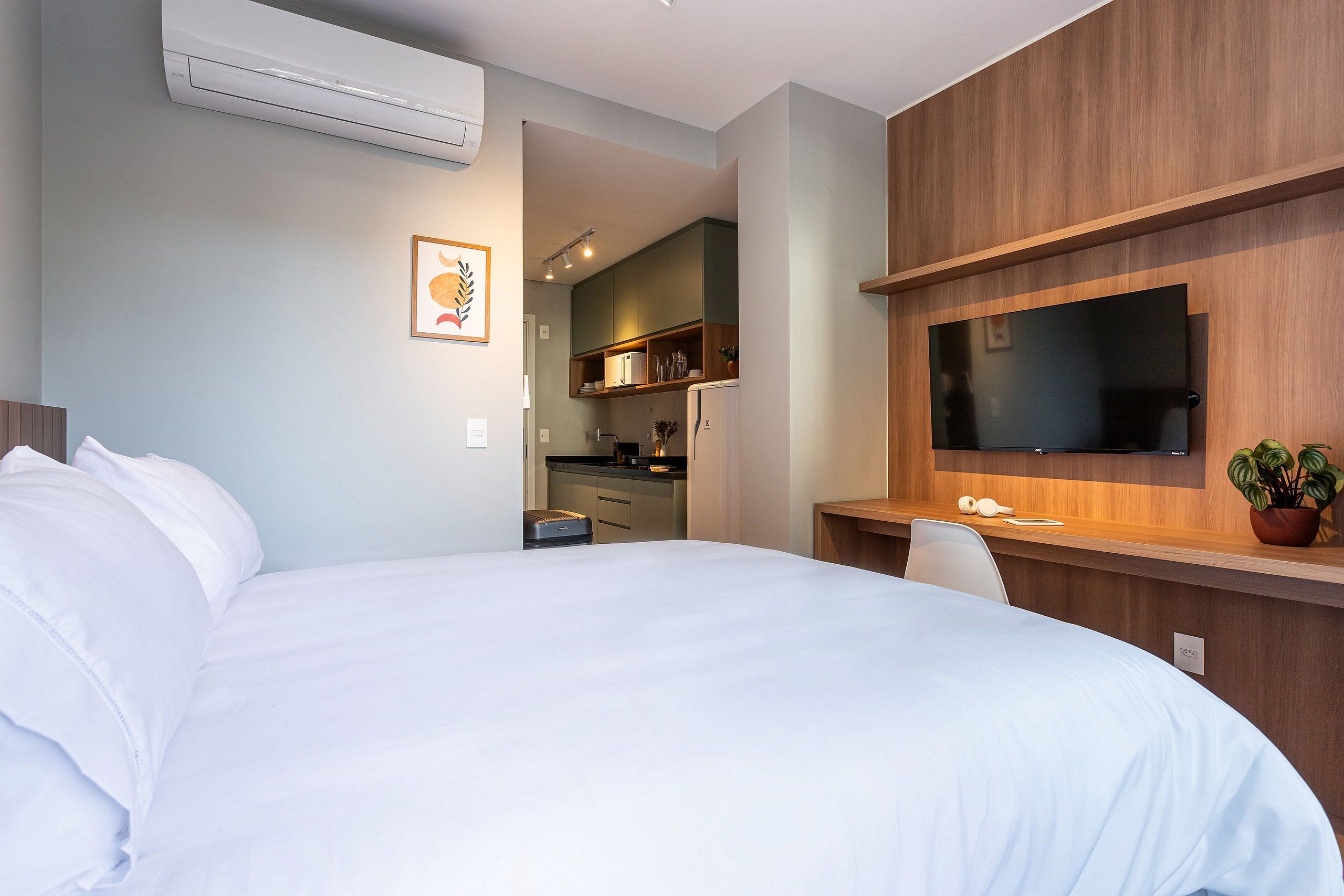A hotel room with a bed, a desk with a flat-screen TV, a potted plant, and a kitchenette in the background.