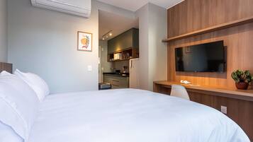 A hotel room with a bed, a desk with a flat-screen TV, a potted plant, and a kitchenette in the background.