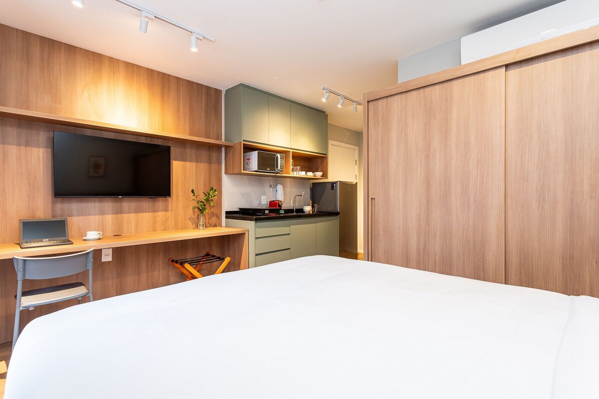 Una habitación de hotel moderna con una cama, un escritorio con una computadora portátil, un televisor, una kitchenette y una puerta corredera.