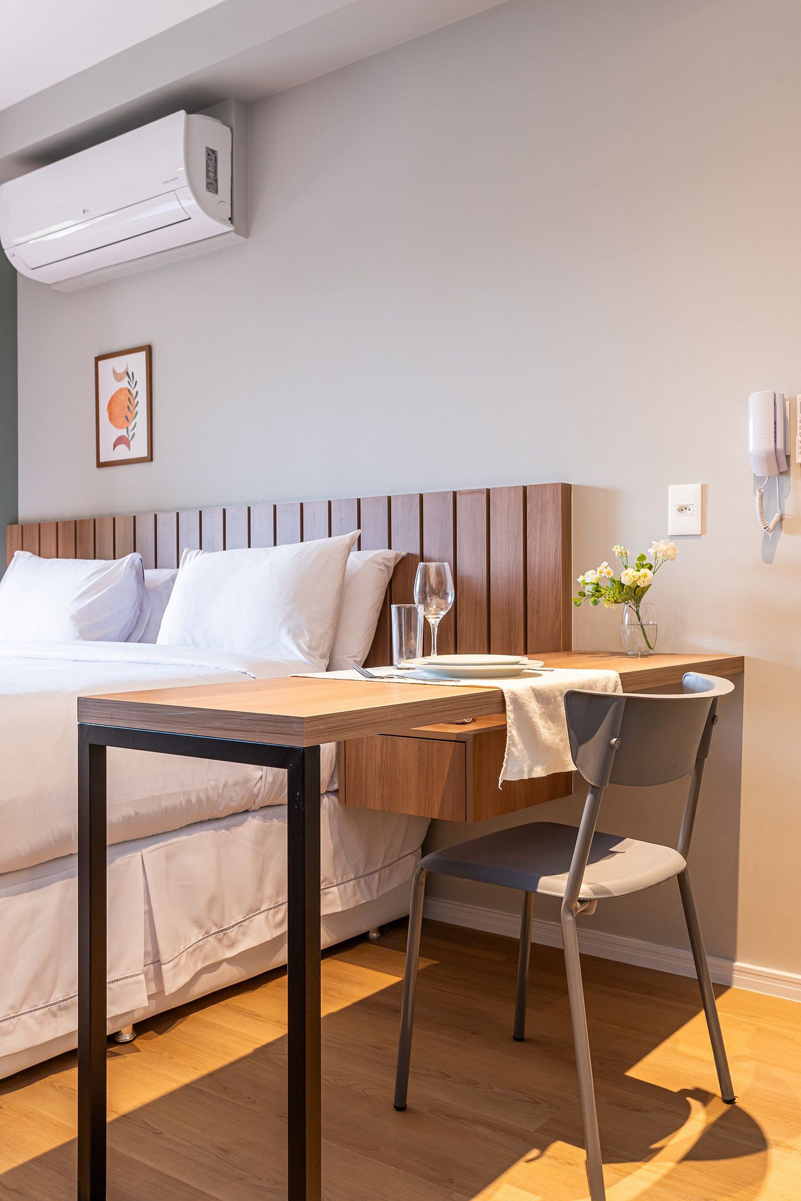 A hotel room with a bed, bedside table, chair, and an air conditioning unit.