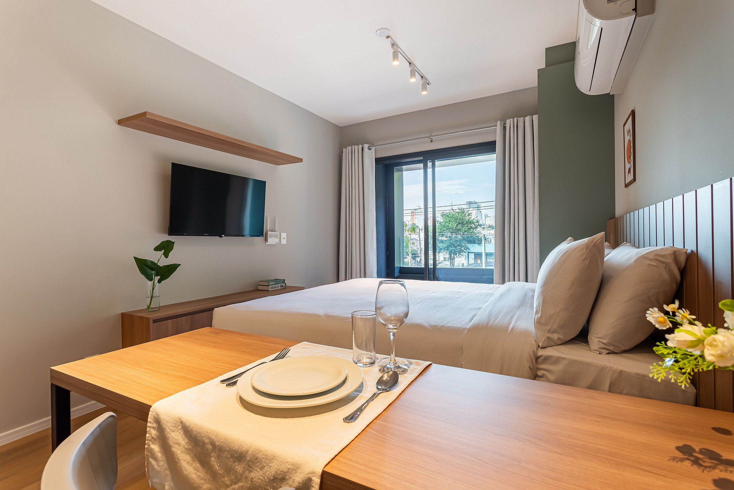 A hotel room with a bed, a TV mounted on the wall, a dining table with a set of plates and cutlery, and a window with curtains.
