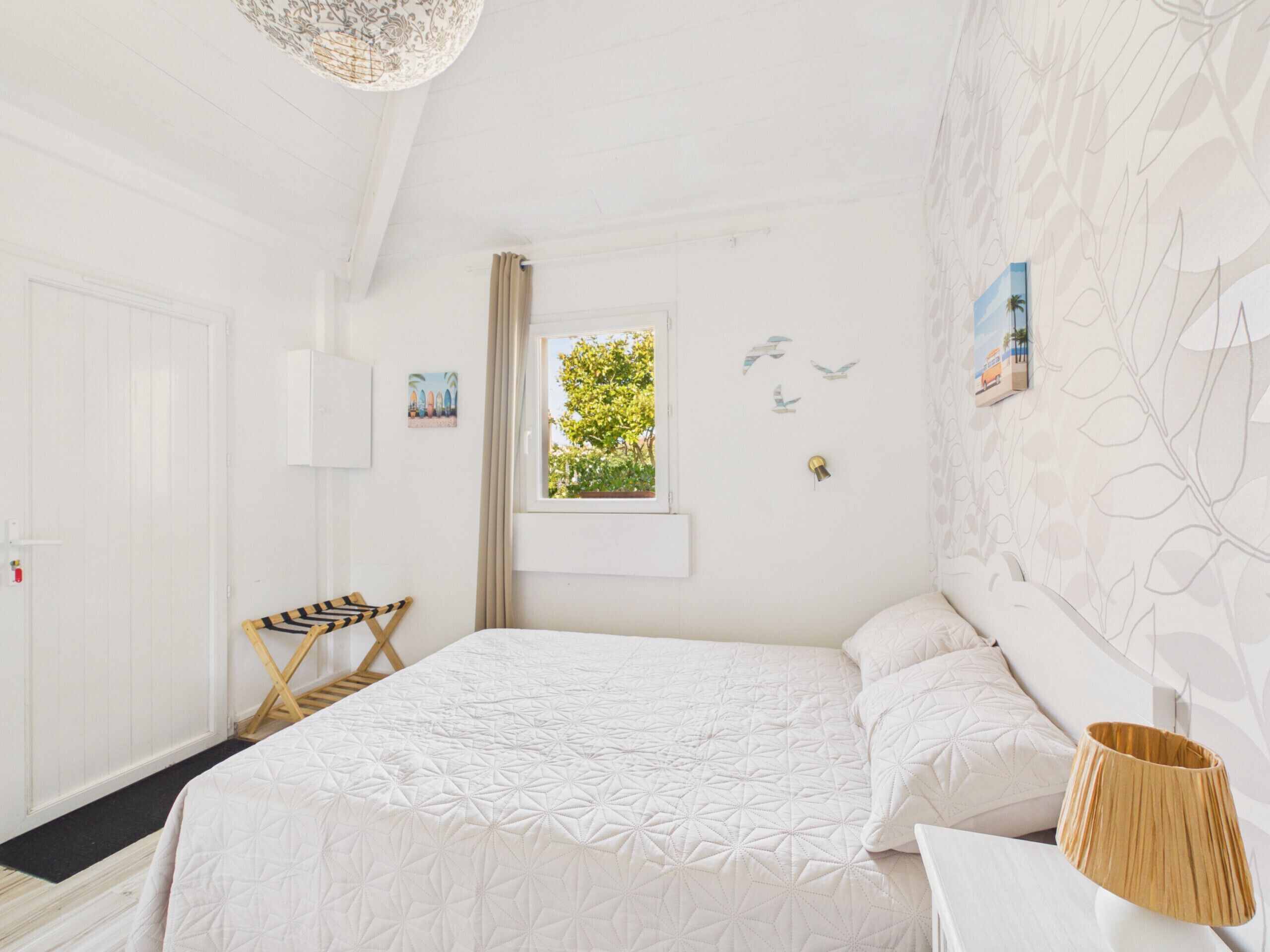 Une pièce blanche avec un lit, une fenêtre donnant sur un paysage verdoyant, une porte et un mur orné d’un motif de feuilles.