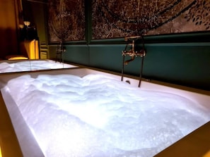 A bathtub with a large, clear water surface, a wall-mounted faucet, and a decorative wall with a tree-like design. - Apartament z Jacuzzi Kraków ART Koletek (Kraków)