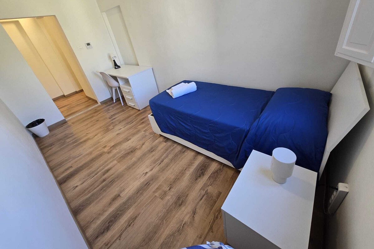 Une petite chambre d’hôtel composée d’une seule pièce, avec un plancher en bois, un lit avec une literie bleue, un bureau avec une chaise et un mur blanc.