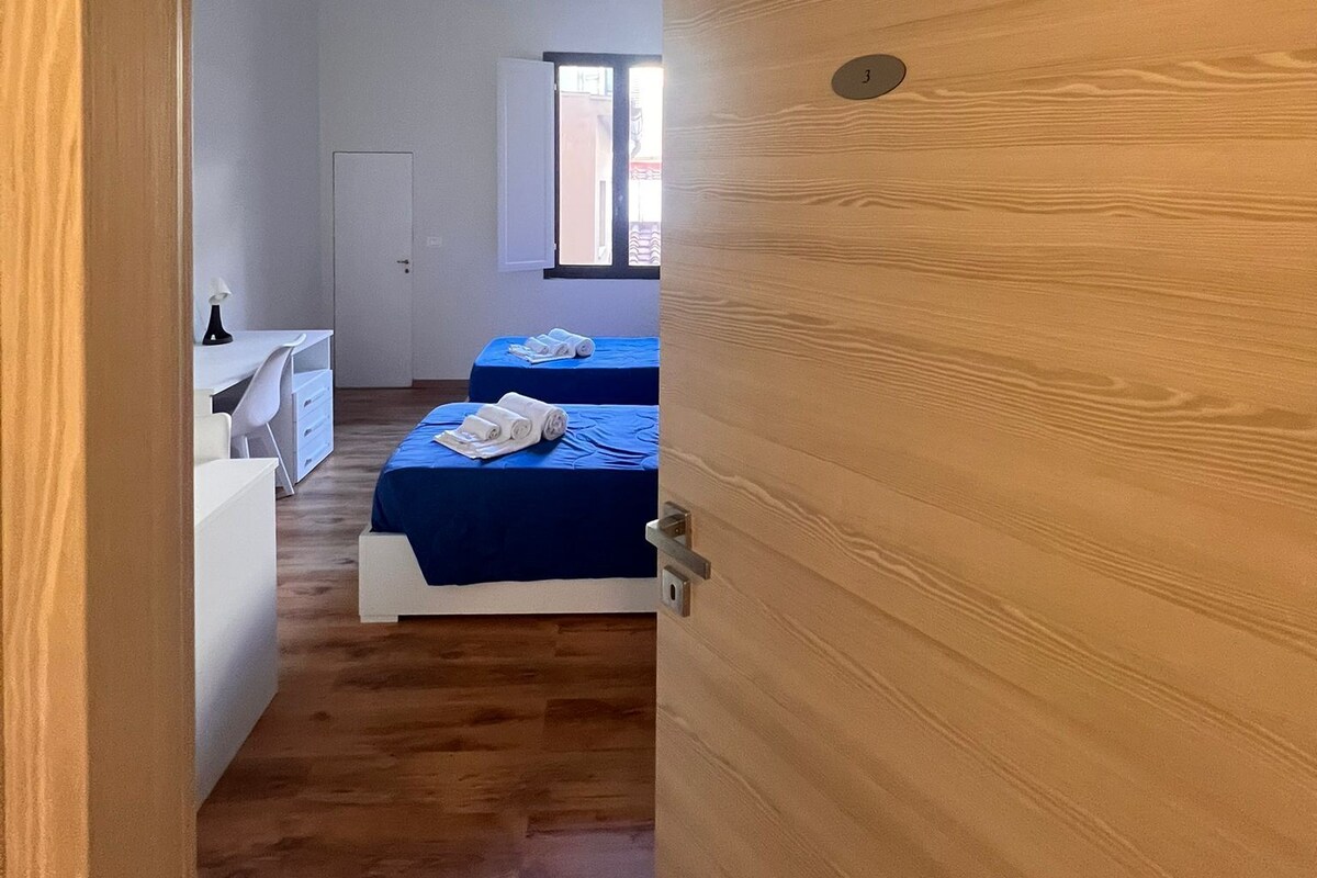 Une pièce avec une porte en bois, un bureau, deux lits avec une literie bleue et une armoire blanche.
